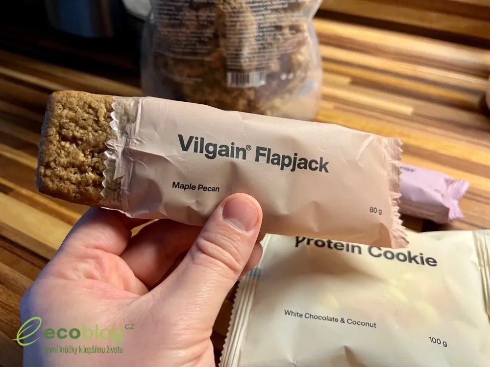 Ruka drží Vilgain Flapjack Maple Pecan s nakousnutým koncem, v pozadí proteinová cookie a granola