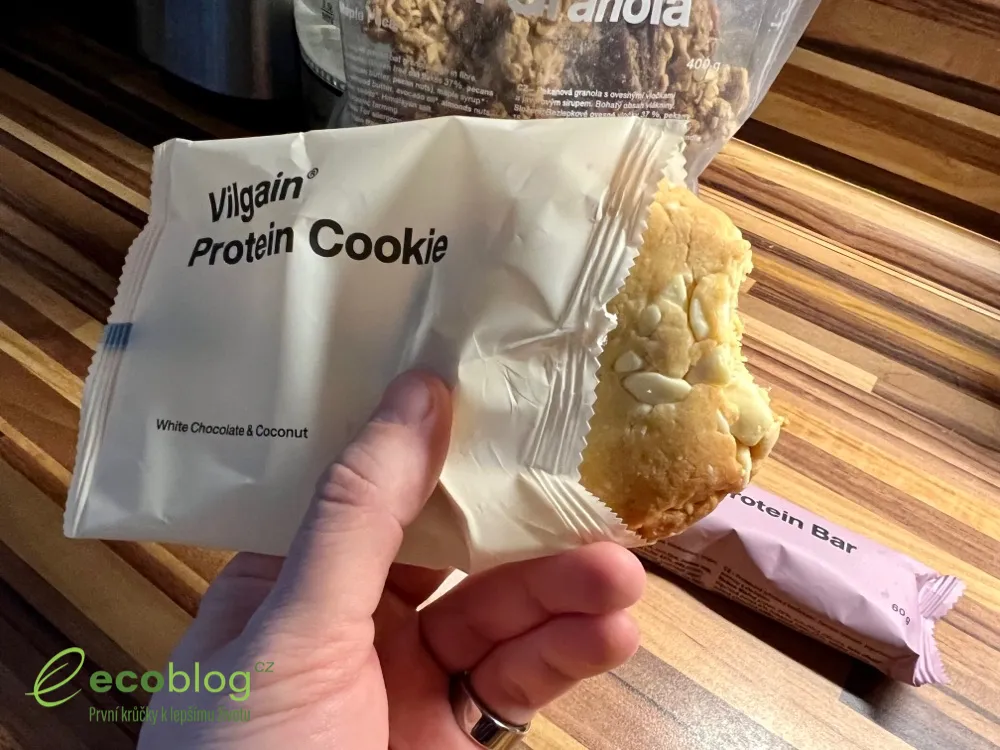 Ruka drží nakousnutou Vilgain Protein Cookie White Chocolate Coconut, v pozadí sáček granoly