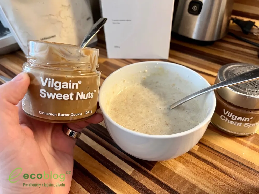 Ruka drží otevřenou sklenici Vilgain Sweet Nuts nad miskou ovesné kaše, vedle stojí Vilgain Cheat Spread