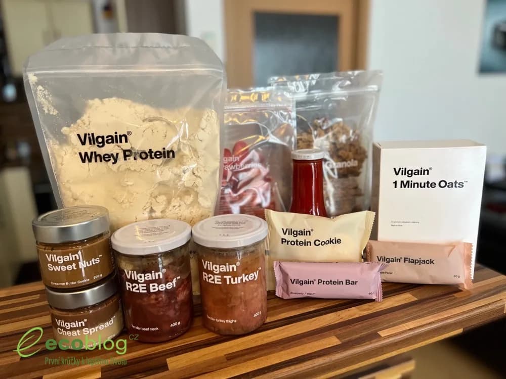Autor recenze sedí u stolu s jedenácti produkty Vilgain, mezi nimi protein, granola, kečup a zavařené maso